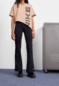 Beige kortärmad skjorta med stor svart "NIKE" tryck, kombinerad med figurnära svarta utsvängda byxor. Modellen har på sig svarta plattformsskor. Grå skåp i bakgrunden.