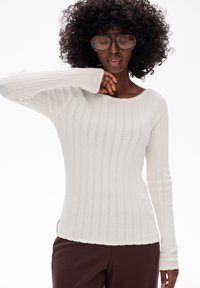 Biała, prążkowana bluzka z długim rękawem, o szerokim dekolcie, z teksturowanym wzorem dzianiny i dopasowanym kroju. W zestawieniu z brązowymi spodniami.