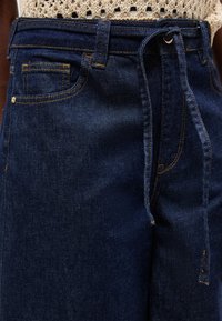 Σκούρα μπλε τζιν παντελόνια με πορτοκαλί ραφές, μπροστινή θηλιά για ζώνη και δεμένη υφασμάτινη ζώνη, φορεμένα με πλεκτό μπεζ τοπ.