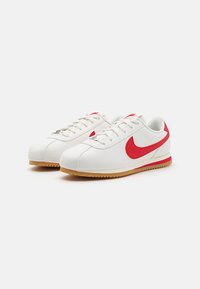 Weiße Sportschuhe mit roten Akzenten, ausgestattet mit einem glatten Leder-Obermaterial, runder Zehenpartie, Schnürsystem und strukturierter Gummisohle.