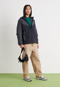 Chaqueta negra y gris con cierre de cremallera y capucha, con bolsillos laterales, llevada sobre una camiseta verde; combinada con pantalones beige y zapatillas grises.