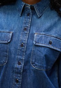 Blå denimskjorte med svarte knapper, to brystlommer med klaff og synlige sømmer på en person med krøllete hår.