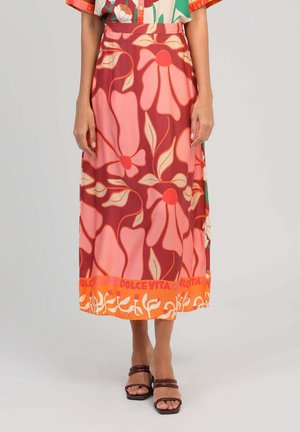 Femme portant une jupe midi avec de grands motifs floraux roses et rouges et un ourlet orange avec texte, associée à des sandales marron à lanières.
