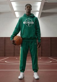 Mand i grøn Celtics-træningsdragt, der holder en basketball og står på en indendørs basketballbane.