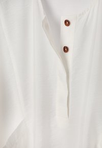 Tissu blanc avec une ouverture en V comportant deux boutons marron sur une patte.