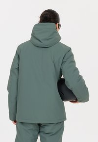 Grön vattentät jacka med hög krage och huva, med en slät textur och håller en svart hjälm i vänster hand.