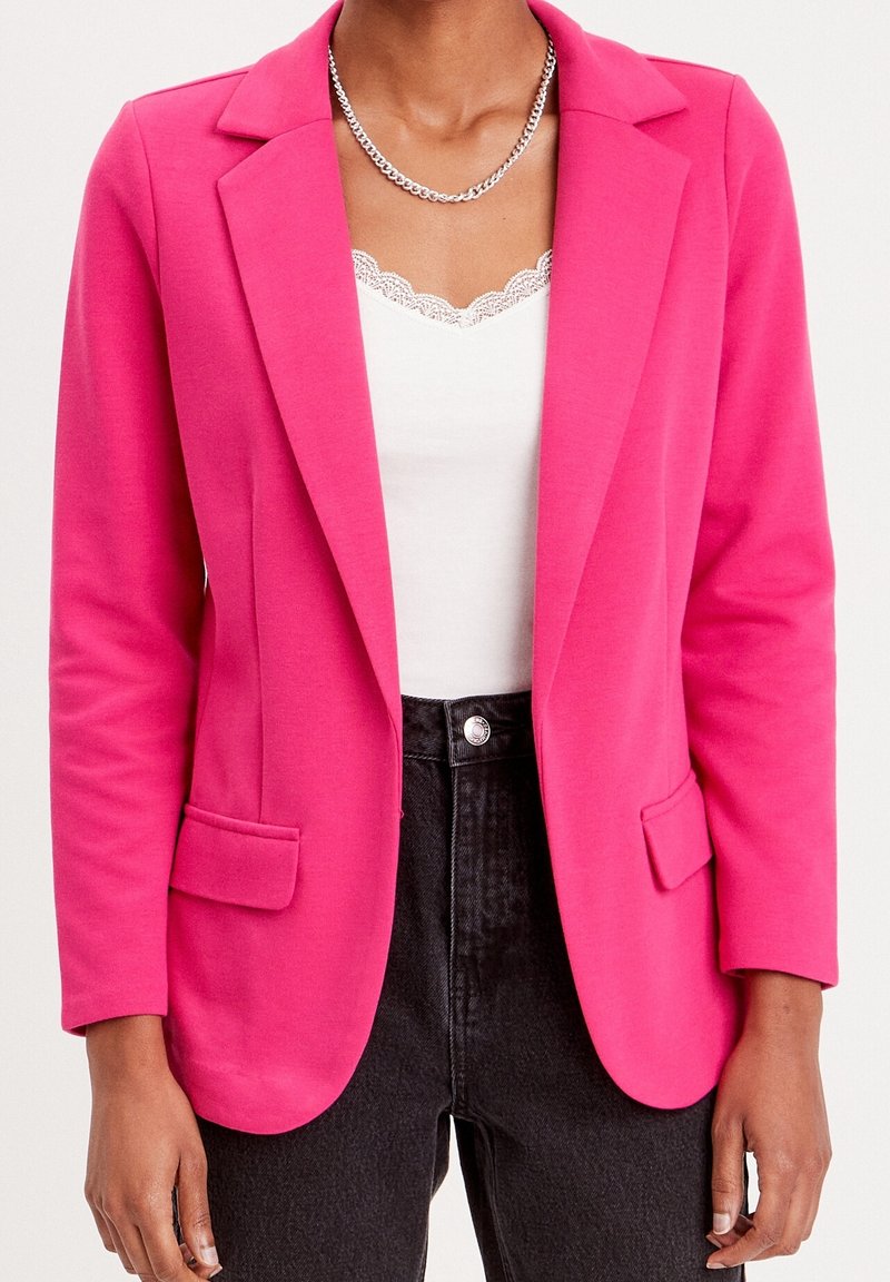 Personne portant un blazer rose vif, un haut blanc à bordure en dentelle, un jean noir taille haute et un collier chaîne argenté.