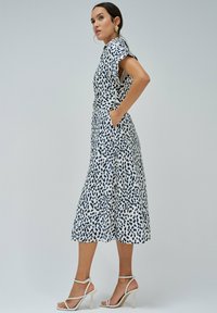 Bílé šaty s námořnickými leopardími vzory, délka do poloviny lýtek, krátké rukávy, detail s páskem, s bočními kapsami. Nosené s bílými sandály na podpatku.