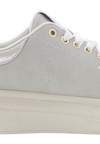 Beige mokkanahka lenkkarit, joissa on kohokuvioitu logo, kultaiset rei'itykset ja valkoiset nauhat. Matala malli, jossa on teksturoitu kumipohja. Minimalistinen estetiikka.