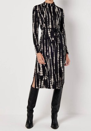 Femme portant une robe midi noire à manches longues avec des rayures verticales tie-dye beige, ceinture à la taille, fente latérale et bottes noires montantes jusqu'au genou.