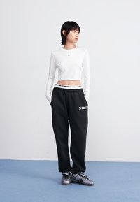 Hvit langermet crop top med en liten svart logo, kombinert med løse svarte joggebukser med en hvit trykt logo. Grå treningssko.