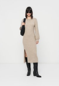Robe en tricot beige avec de longues manches ballon et une fente sur le côté, associée à des bottes noires jusqu'aux genoux et un sac à épaule noir.