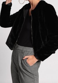 Black faux fur jacket with a zip closure, featuring a high neck and long sleeves. Worn over fitted black top and houndstooth patterned trousers.
