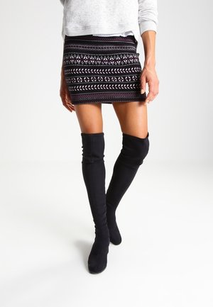 Kobieta ubrana w czarną mini spódnicę w wzory, jasnoszary sweter i czarne kozaki za kolano stojąca na białym tle.