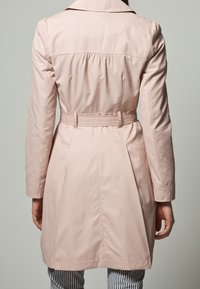 Ljusrosa trenchcoat med insydd midja, långa ärmar och krage. Har en slät textur och subtila rynkade detaljer på ryggen.