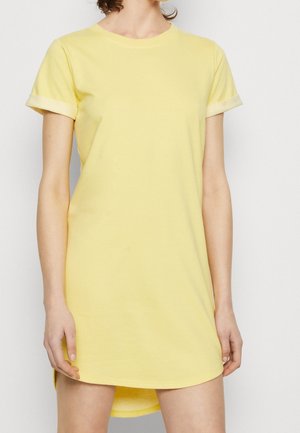 Žena v krátkém žlutém šatu s ohrnutými rukávy stojí před jednobarevným světlým pozadím, ruce volně podél těla.