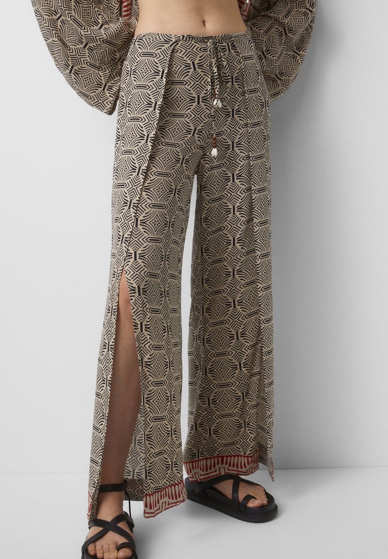 Pantalon beige à jambes larges avec motifs géométriques noirs, fentes latérales sur les jambes, accents de bordure rouge, taille à cordon avec perles, porté avec des sandales noires.