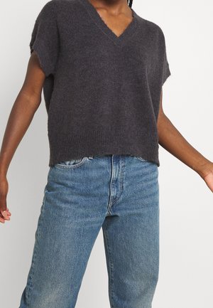 Personne portant un pull ample gris foncé à manches courtes et col en V, associé à un jean en denim bleu taille haute, sur un fond blanc.