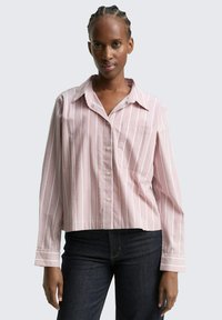 Kvinna med flätat hår som bär en rosa och vit randig skjorta med knappar och mörka jeans, stående mot en ljus enfärgad bakgrund.