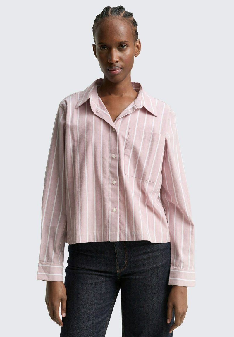 Kvinna med flätat hår som bär en rosa och vit randig skjorta med knappar och mörka jeans, stående mot en ljus enfärgad bakgrund.