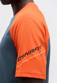 Κοντινό πλάνο ενός ατόμου που φοράει γκρι και πορτοκαλί αθλητική ένδυση με την επιγραφή "DYNAFIT" στο πορτοκαλί μανίκι.