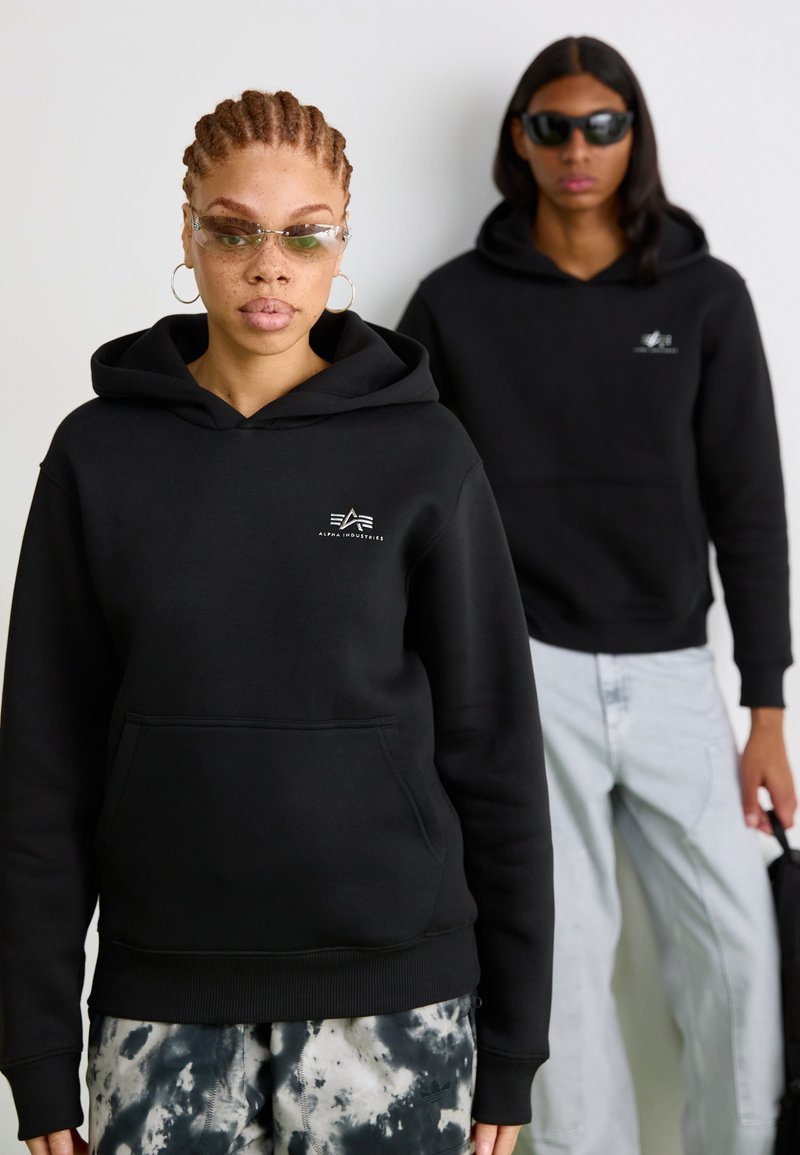 Felpa nera con cappuccio, tasca frontale e logo bianco sul petto. Il modello indossa anche occhiali da sole trasparenti, abbinati a pantaloni tie-dye.