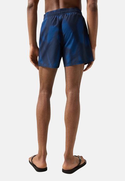 Widok z tyłu osoby noszącej niebieskie kąpielówki i czarne klapki, stojącej na tle jednolitego jasnego tła.