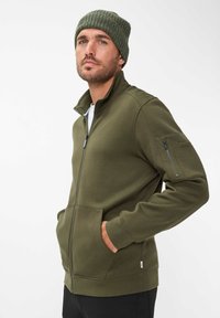 Giacca zip-up verde oliva con colletto alto, due tasche frontali e un ulteriore taschino con zip sulla manica sinistra. Tessuto dalla consistenza morbida.