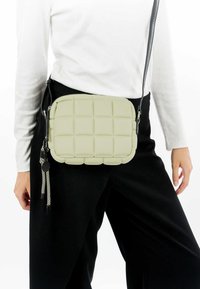 Bolso de bandolera acolchado en silicona verde claro con un diseño de cuadros, que cuenta con una correa negra desmontable con flecos decorativos.