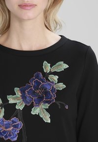 Svart topp med rund halsringning, prydd med flerfärgad blomstersbroderi i lila, grönt och ljust brunt, vilket tillför textur och detaljer.