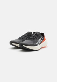Black and gray athletic shoes with a textured mesh upper, prominent white "TERREX" branding, and a vibrant orange heel accent.
