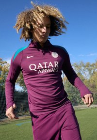 Violetti urheiluasu, jossa on pitkät hihat, vetoketjukaulus ja turkoosit yksityiskohdat. Takissa on Qatar Airwaysin logo ja Pariisin tunnus rinnassa.