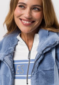 Hellblaue Kunstpelzjacke mit durchgehendem Reißverschluss, ausgestattet mit einem hohen Kragen und kontrastierenden blauen Akzenten. Strukturierter Stoff mit leichtem Glanz.