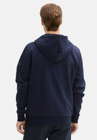 Navyblauwe hoodie met een textuurafwerking, voorzien van een grote capuchon, lange mouwen en ribgebreide boorden. Onderschatte logo detail op de arm.