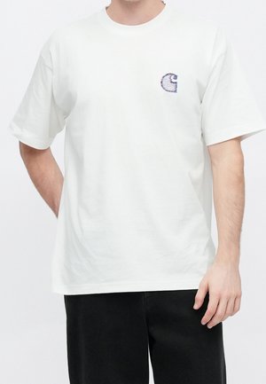 Homme portant un T-shirt blanc à manches courtes avec un petit logo brodé sur la poitrine gauche, associé à un pantalon noir.
