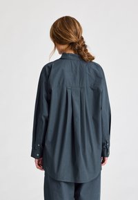 Camicia oversize in misto cotone, grigio-blu scuro, con una piega sul retro e polsini con bottoni. Texture liscia con un design di colletto classico.