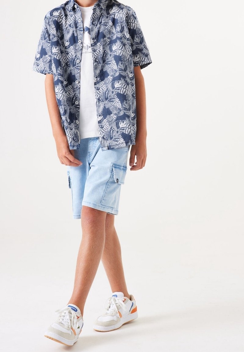 Enfant portant une chemise bleue à manches courtes à motifs, boutonnée, sur un t-shirt blanc, un short cargo bleu clair et des baskets blanches avec des accents orange.