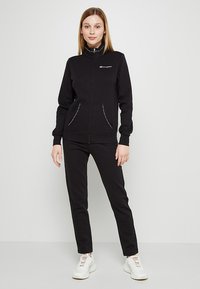 Sudadera negra con cremallera y cuello alto, con dos bolsillos frontales. Combinada con pantalones negros a juego y zapatillas blancas. Textura suave.