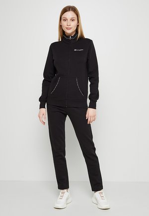 Sudadera negra con cremallera y cuello alto, con dos bolsillos frontales. Combinada con pantalones negros a juego y zapatillas blancas. Textura suave.