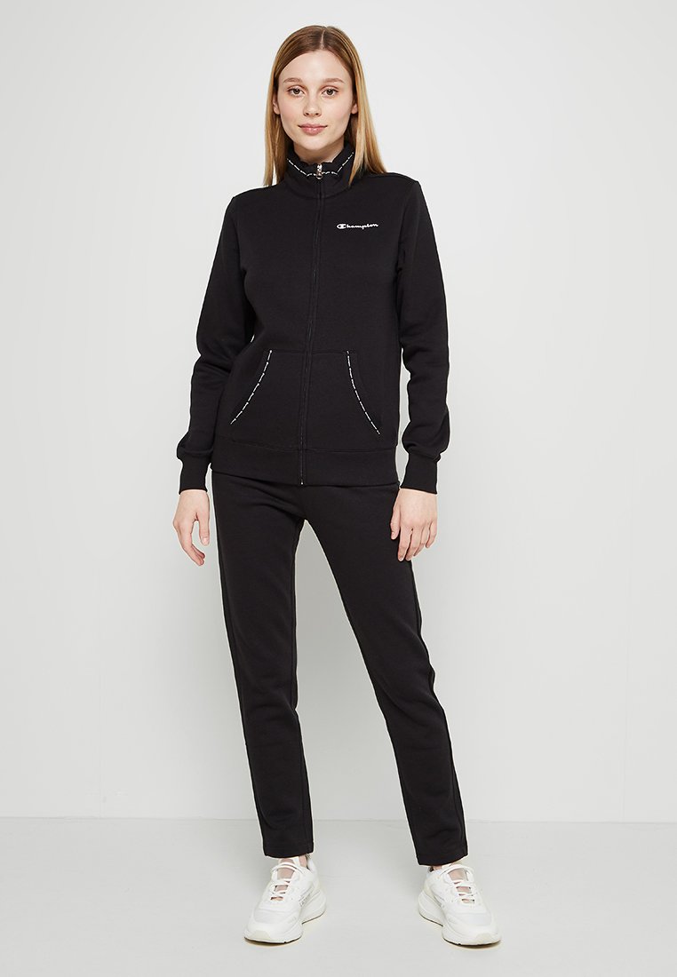 Sudadera negra con cremallera y cuello alto, con dos bolsillos frontales. Combinada con pantalones negros a juego y zapatillas blancas. Textura suave.