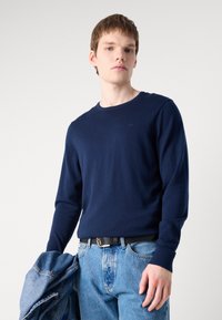 Pulover navy blue cu guler rotund, din material moale, design ajustat. Modelul ține o jachetă din denim, purtând blugi albaștri deschiși cu un cureaua neagră.