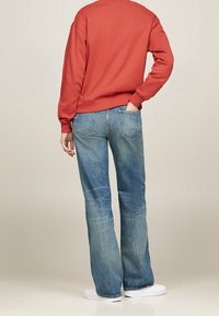 Roter Sweatshirt mit lockerer Passform, kombiniert mit ausgewaschenen blauen Schlagjeans und weißen Turnschuhen. Die Jeans haben Gesäßtaschen und eine helle Waschung.