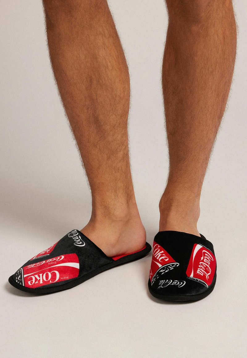 Černé a červené nazouvací pantofle s logem Coca-Cola a designy plechovek, vyrobené z měkké látky s plochou podrážkou.
