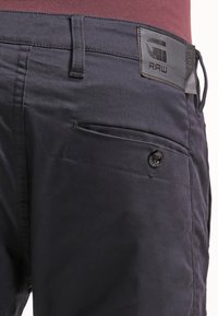 Mörkblå byxor med texturerat tyg, featuring en knappstängning, sidofickor och ett svart logopatch på midjan.