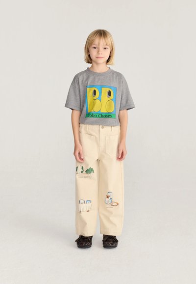 Enfant aux cheveux blonds debout, portant un t-shirt gris avec un motif coloré et un pantalon beige avec des dessins imprimés ludiques.