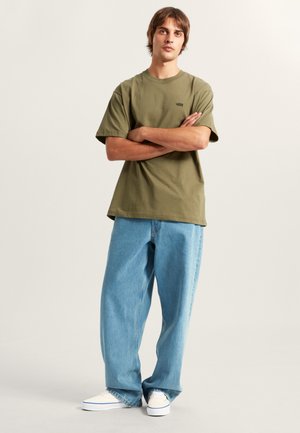 Joven con los brazos cruzados, lleva una camiseta Vans verde oliva, vaqueros sueltos azul claro y zapatillas blancas sobre un fondo liso.