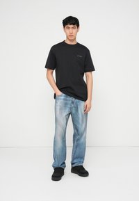 Νεαρός άνδρας που στέκεται φορώντας μαύρο μπλουζάκι, ξεθωριασμένο μπλε τζιν και μαύρα αθλητικά παπούτσια μπροστά σε απλό λευκό φόντο.