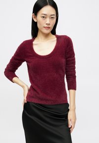 Egyenes fekete hajú nő bolyhos, bordó színű, hosszú ujjú felsőt visel, és egy fekete selyem szoknyával pózol, egyik kezét csípőre téve, fehér háttér előtt.