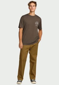 Jovem de pé com uma mão no bolso, vestindo uma t-shirt gráfica castanha escura, calças castanhas claras e ténis preto e branco.