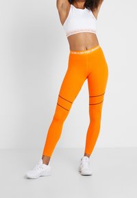 Sportowy stanik Calvin Klein Performance oraz jasnopomarańczowe legginsy charakteryzują się kontrastowymi czarnymi paskami i gładką elastyczną talią.